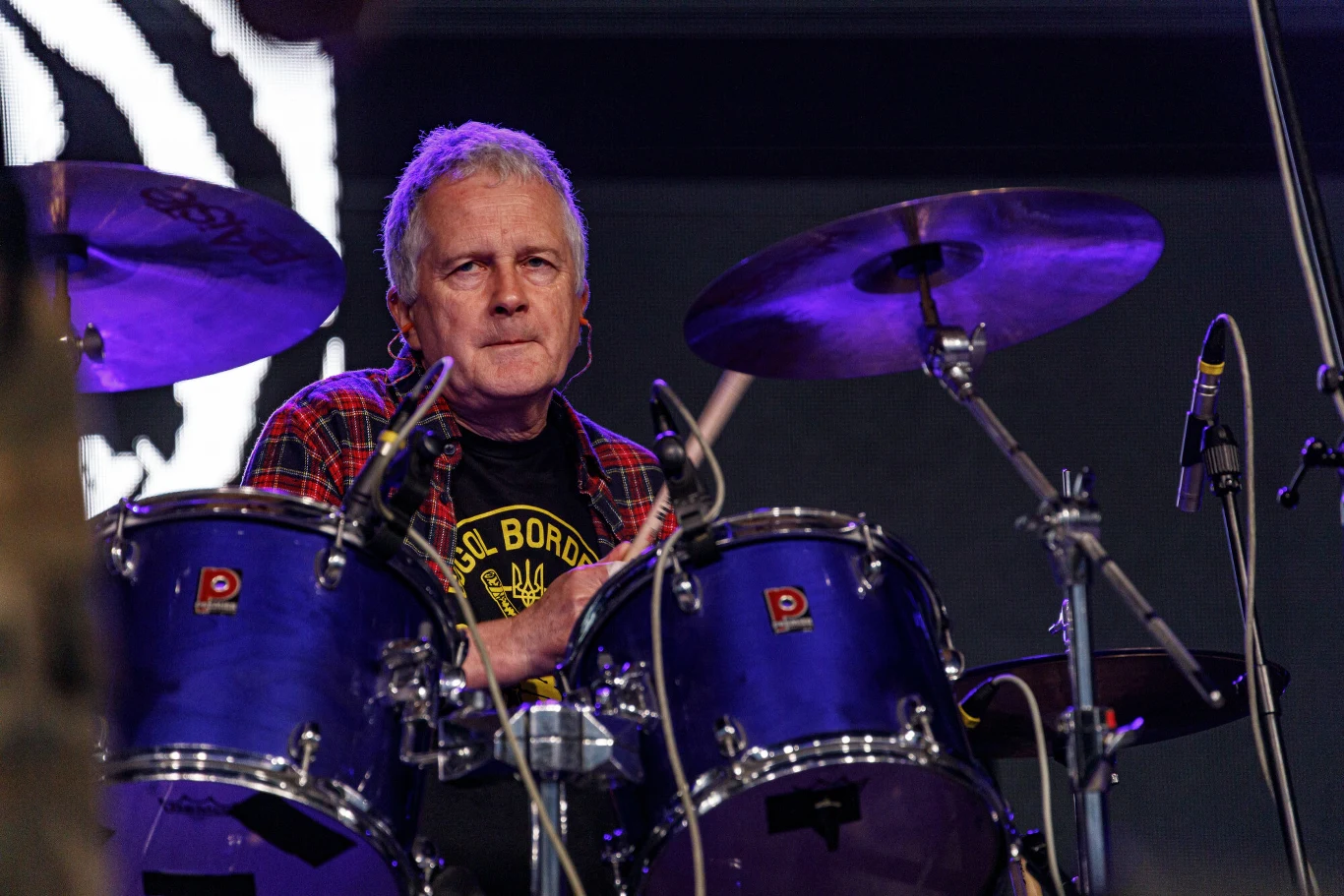 Krzysztof Grabowski (Dezerter) Starszy mężczyzna gra na perkusji podczas koncertu, skupiony na swojej grze, w tle widoczne elementy sceny oraz oświetlenie koncertowe.