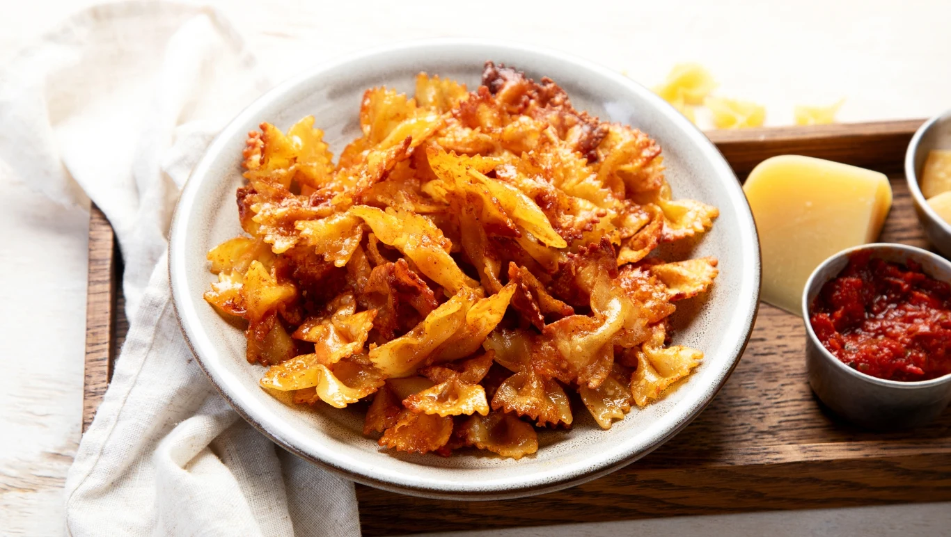 Talerz chrupiącego makaronu farfalle, podanego na białym talerzu z dodatkiem kawałka sera oraz miseczki z czerwonym sosem, ustawionych na drewnianej desce.