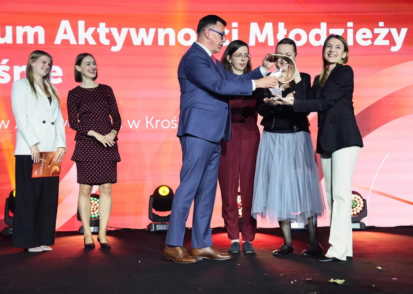 Nagroda Przewroty – edukacja to powód do dumy Grupa osób ubranych w eleganckie stroje stoi na scenie, część z nich trzyma szklaną statuetkę przekazywaną sobie nawzajem. W tle czerwona ścianka z napisem związanym z aktywnością młodzieży.