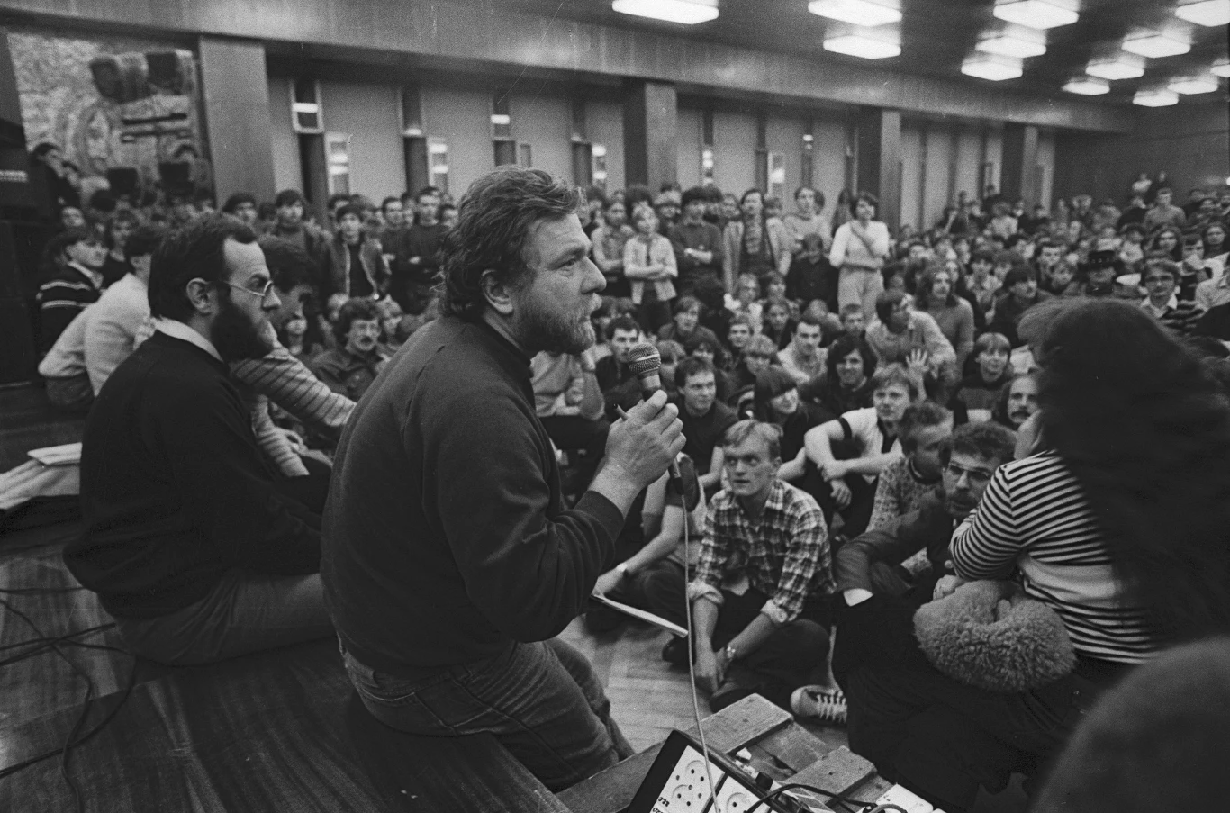 Piotr Kaczkowski podczas Rockowiska w Łodzi w 1983 r. Mężczyzna z mikrofonem w ręku przemawia do licznie zgromadzonego tłumu siedzącego na podłodze w dużej sali, wokół panuje atmosfera skupienia i zainteresowania; na pierwszym planie widoczne są osoby prowadzące spotkanie, w tle zgromadzenie ludzie.
