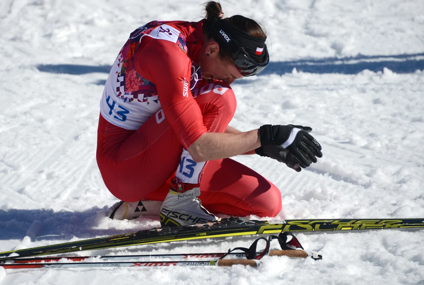 Justyna Kowalczyk po wywalczeniu złotego medalu na igrzyskach w Sochi, 2014 Zawodniczka w czerwonym stroju narciarskim klęczy na śniegu ze spuszczoną głową, obok leżą narty biegowe oraz kijki. Na twarzy widoczne napięcie i zmęczenie po wyścigu.