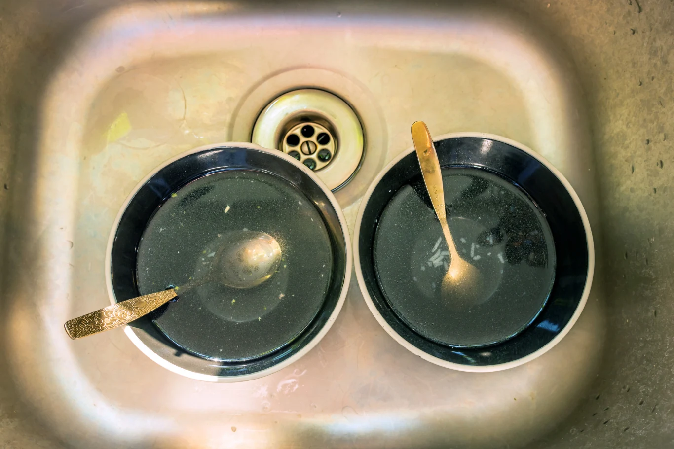 Dwie miski z brudną wodą i łyżkami znajdują się w zlewie kuchennym, wokół nich widać resztki jedzenia, a dno zlewu jest lekko zabrudzone.