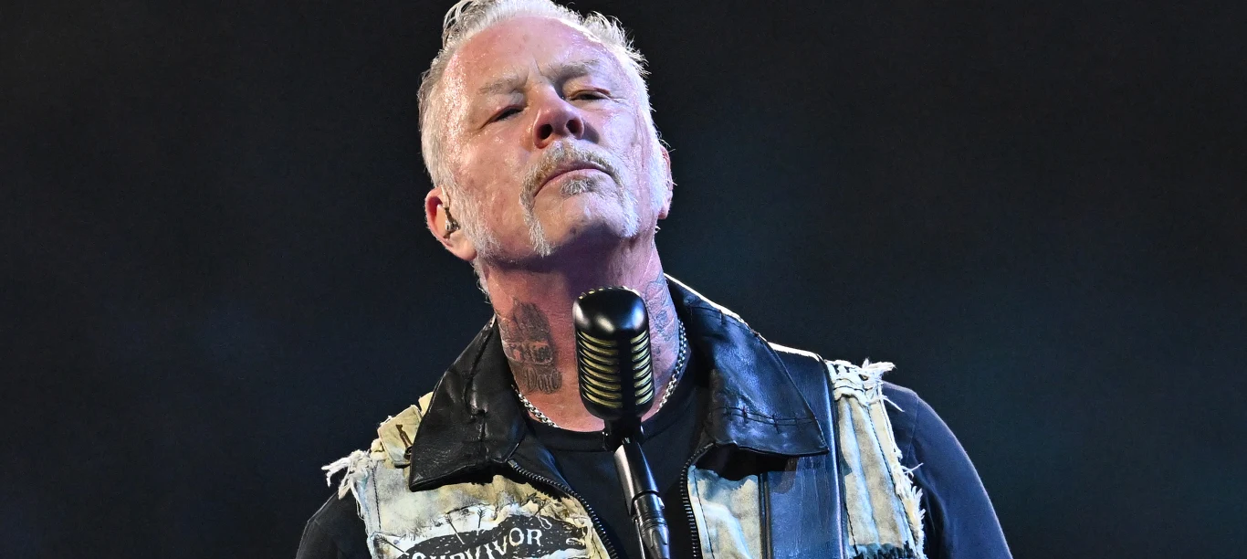 James Hetfield Mężczyzna z siwiejącymi włosami i wąsami, ubrany w kamizelkę z naszywką, stoi przed mikrofonem podczas występu na scenie, patrząc przed siebie z poważnym wyrazem twarzy.