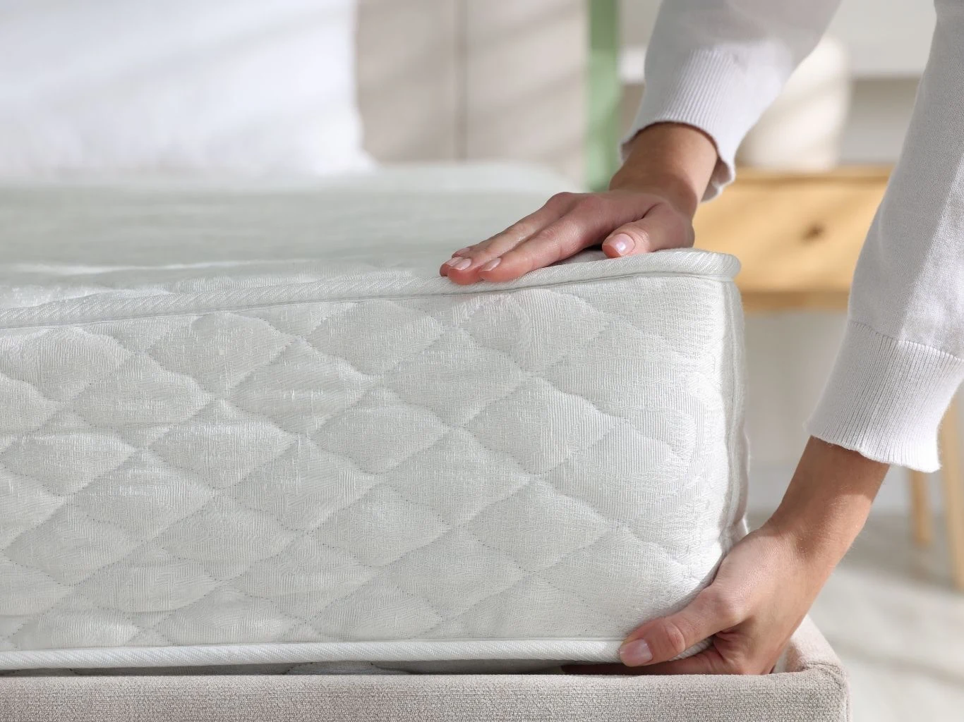 Ręce ustawiające materac na łóżku, materac o jasnym pikowanym pokrowcu na tle rozmazanego wnętrza sypialni