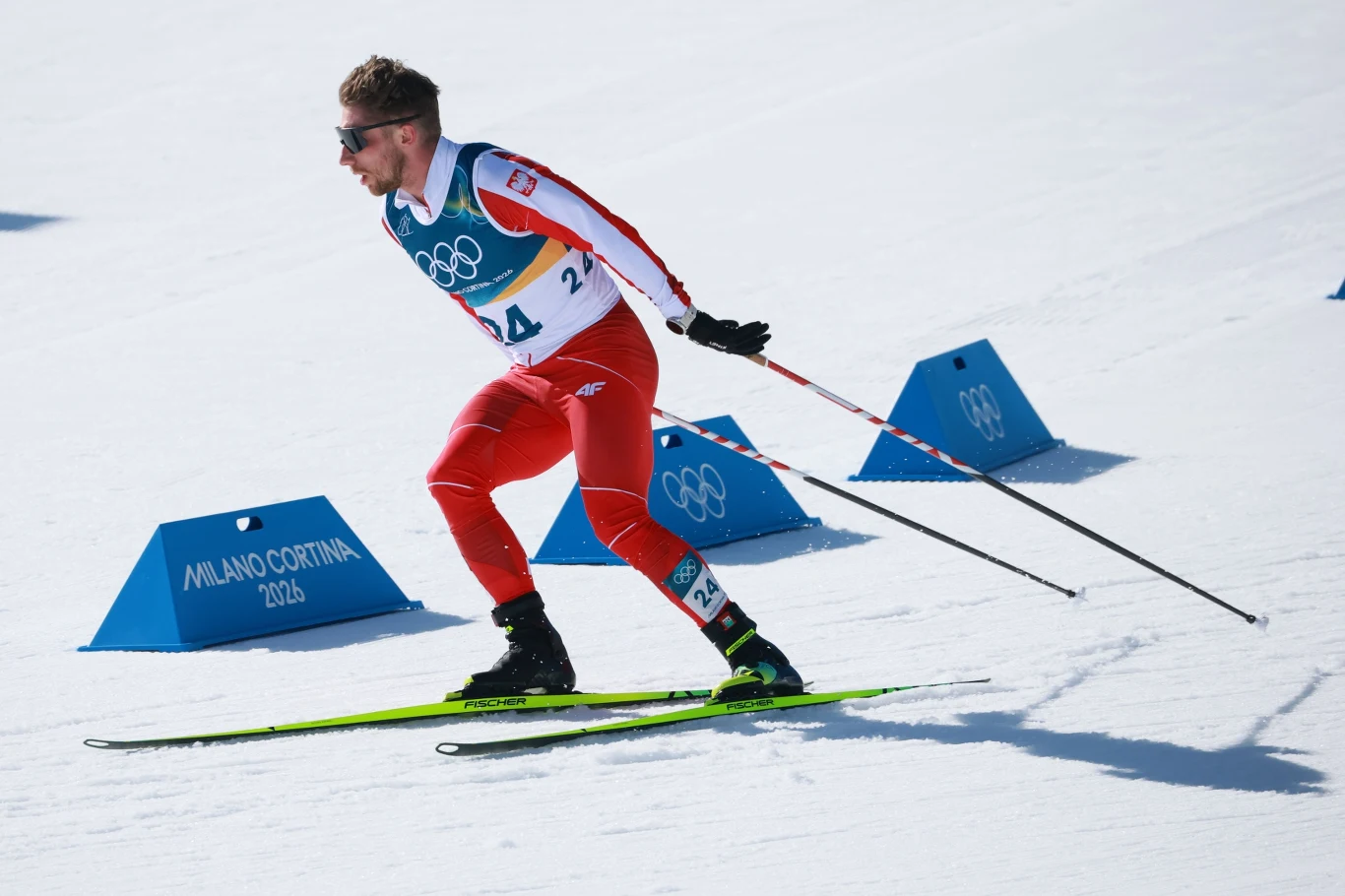 Narciarz biegowy w stroju olimpijskim rywalizuje na ośnieżonej trasie podczas zawodów, w tle niebieskie znaczniki z napisem Milano Cortina 2026.