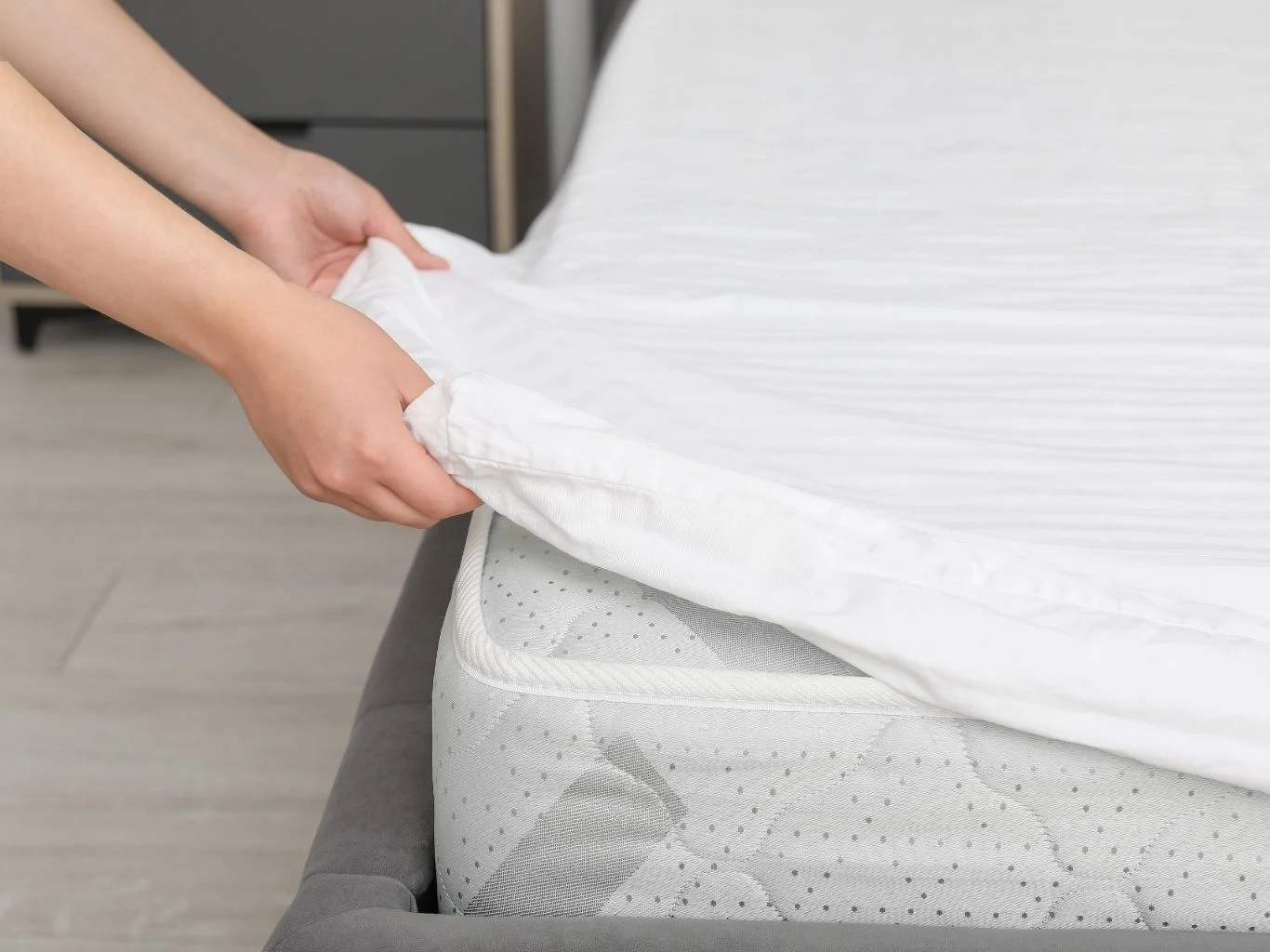 Ręce nakładające świeżą, białą prześcieradło na materac łóżka, widoczna gładka powierzchnia materaca oraz fragmenty łóżka i podłogi.