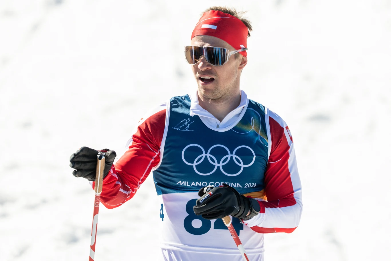 Narciarz w czerwonym stroju sportowym, z opaską na głowie i okularami przeciwsłonecznymi, bierze udział w zawodach olimpijskich w biegu narciarskim. Trzyma kijki narciarskie, na piersi widnieją symbole igrzysk olimpijskich oraz numer startowy.