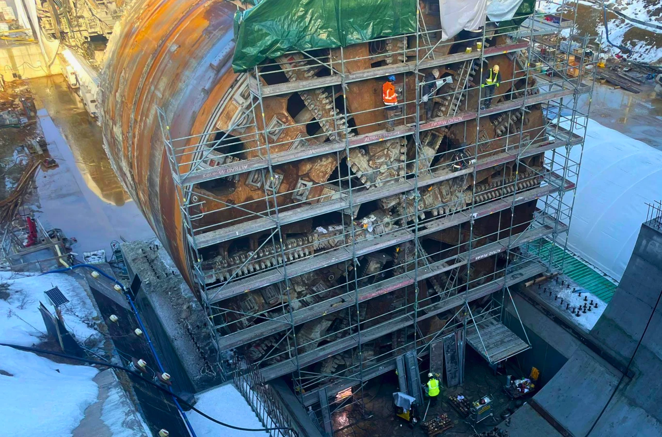 "Karpatka" zakończyła obrót. Gigant ważący 4,4 tys. ton na S19 gotowy. Gigantyczna głowica tarczy drążącej tunel otoczona rusztowaniami, na których pracują robotnicy w kaskach ochronnych. Widoczne są elementy mechaniczne głowicy oraz mokra, błotnista powierzchnia terenu budowy.