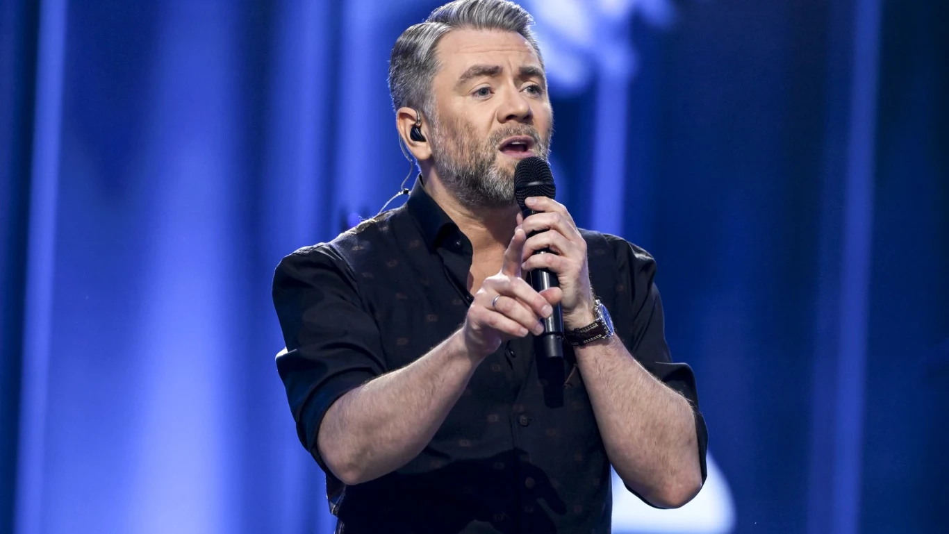 Kuba Badach Mężczyzna z siwiejącymi włosami i zarostem śpiewa do mikrofonu na scenie, ubrany w ciemną koszulę, w tle niebieskie światła i zamazane sylwetki stanowiące tło koncertowe.