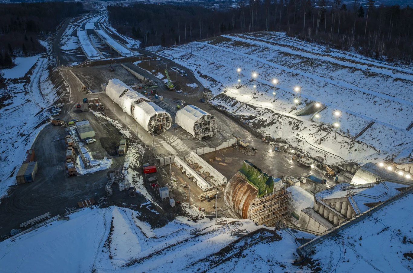 "Karpatka" zakończyła kluczowy manewr. Rozległy plac budowy tunelu w zimowej scenerii z widocznymi dużymi maszynami tunelującymi, oświetlonymi reflektorami, rozkopaną ziemią oraz maszynami budowlanymi, otoczony śnieżnymi zboczami i lasem w tle.