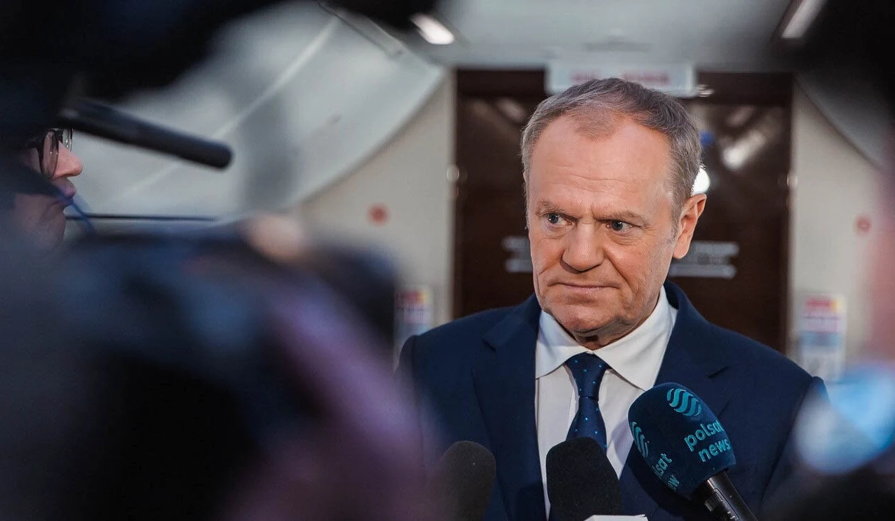 Donald Tusk odniósł się do kwestii sporu politycznego w Polsce wokół programu SAFE Mężczyzna w garniturze i krawacie stoi w otoczeniu mikrofonów oraz kamer dziennikarzy podczas wypowiedzi prasowej w nowoczesnym wnętrzu.