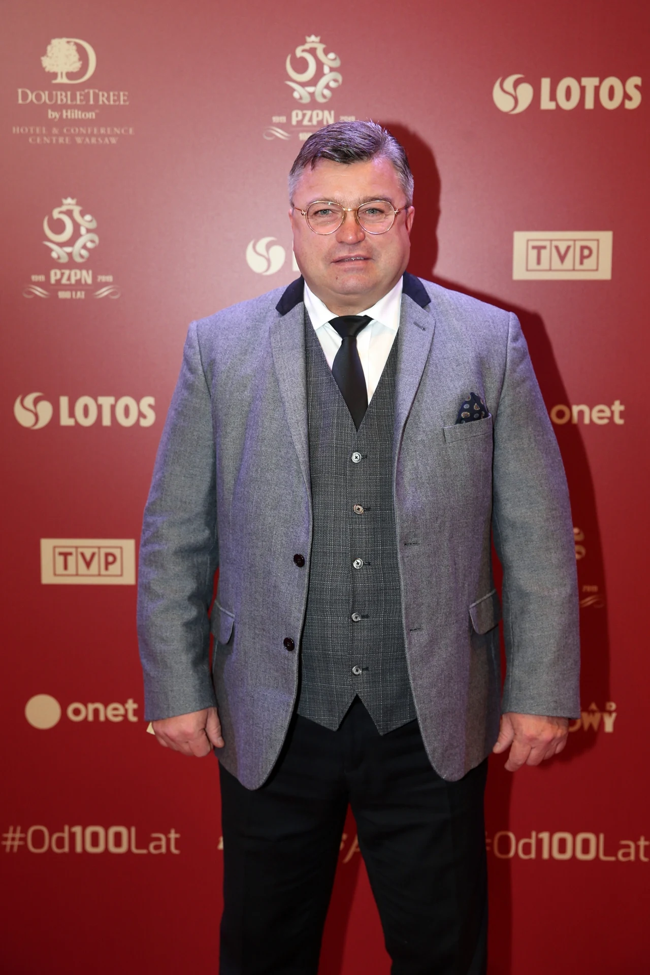 Roman Kosecki Mężczyzna w szarym garniturze i czarnej kamizelce stoi na tle czerwonej ścianki z logotypami sponsorów oraz partnerów wydarzenia.