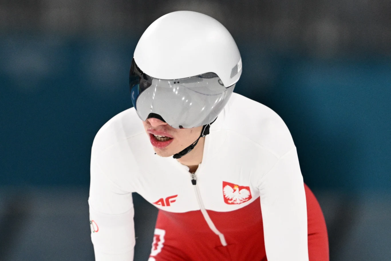 Władimir Semirunnij Zawodnik w stroju do łyżwiarstwa szybkiego w barwach Polski, z kaskiem i okularami ochronnymi, pochylony w pozycji startowej na lodowisku.