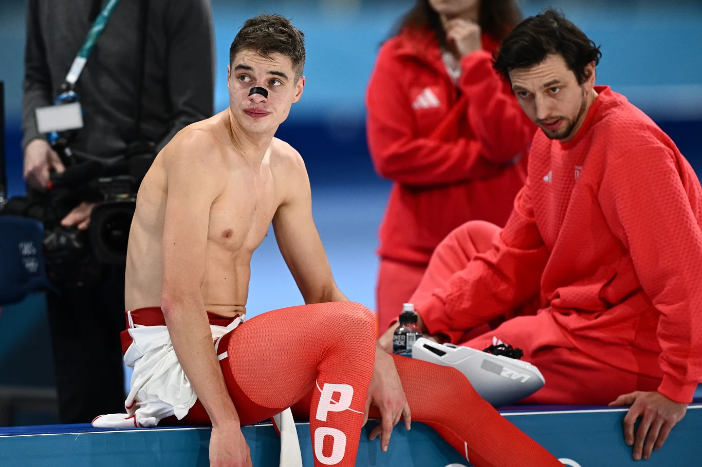 Sportowiec w czerwono-białych legginsach i białym ręczniku siedzi na ławce po zawodach, obok niego druga osoba w czerwonym dresie, w tle rozmazana sylwetka kobiety i sprzęt sportowy.