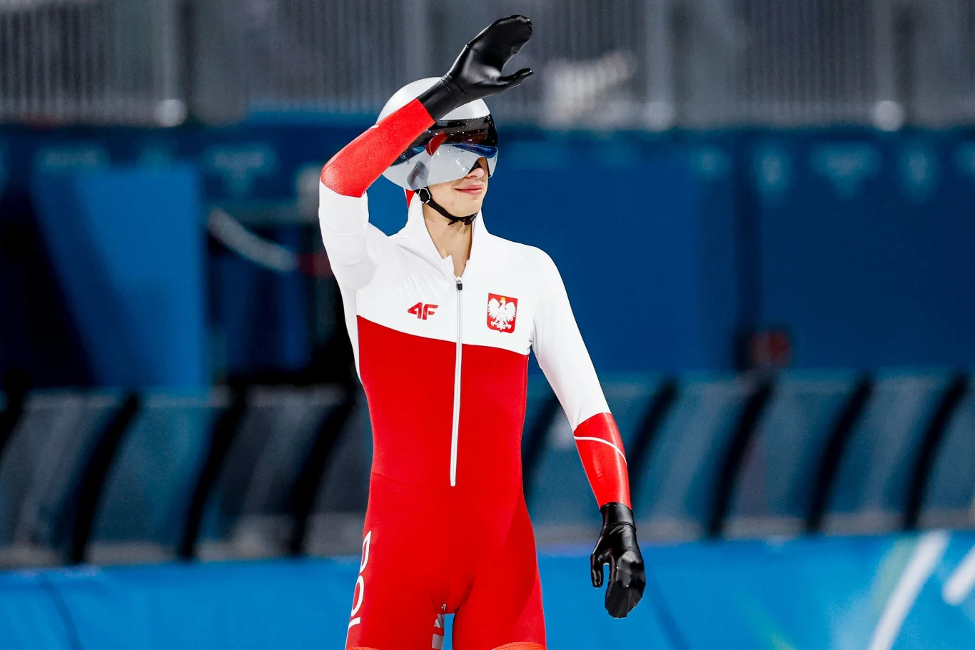 Zawodnik w biało-czerwonym stroju reprezentacji Polski do łyżwiarstwa szybkiego stoi na lodowisku z uniesioną ręką w geście osłaniającym twarz, w tle widoczne puste trybuny