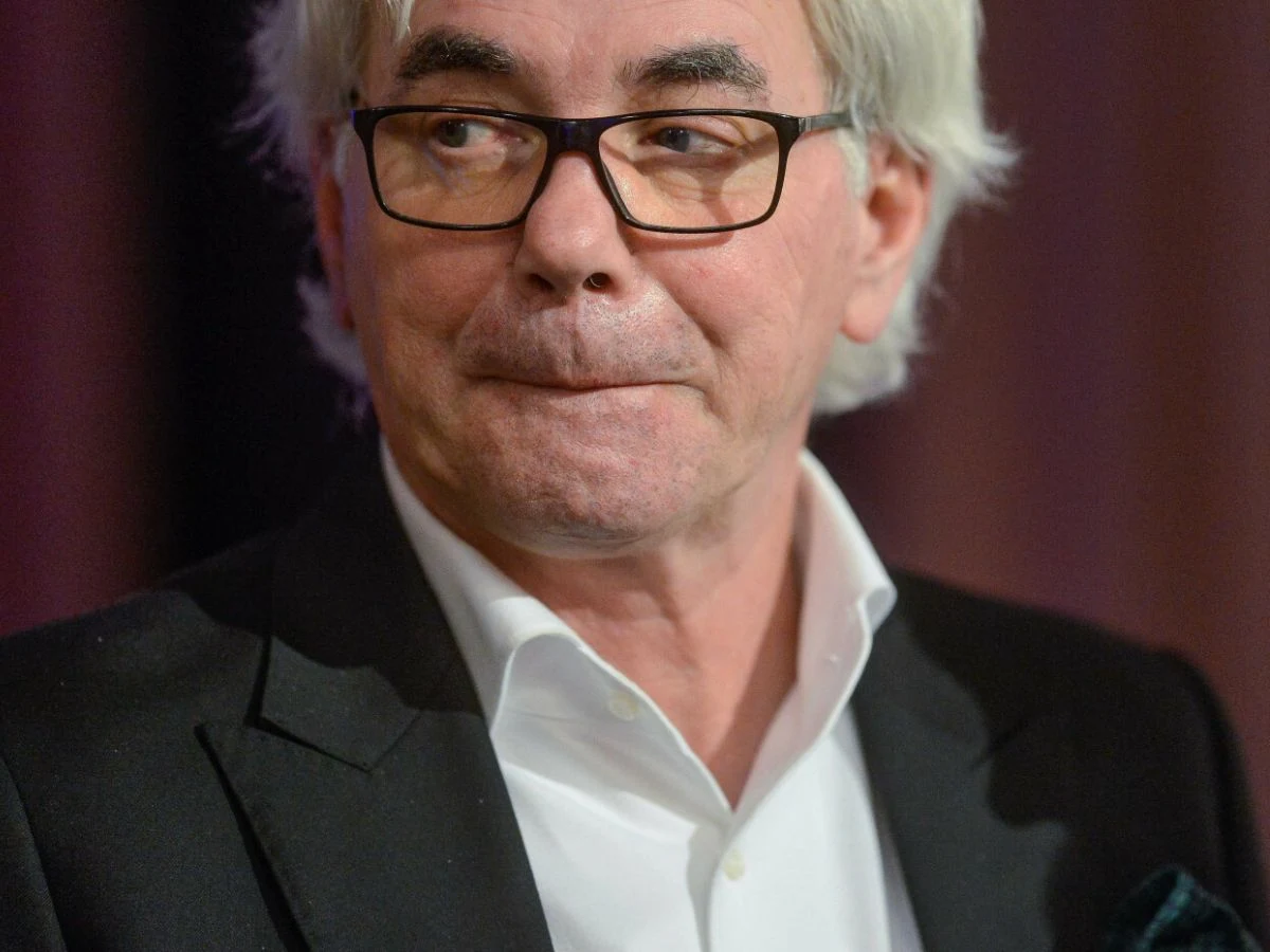 Robert Janowski Starszy mężczyzna o siwych włosach i okularach w eleganckim garniturze i białej koszuli, patrzący w bok, o poważnym wyrazie twarzy. Tło rozmyte, w odcieniach fioletu.