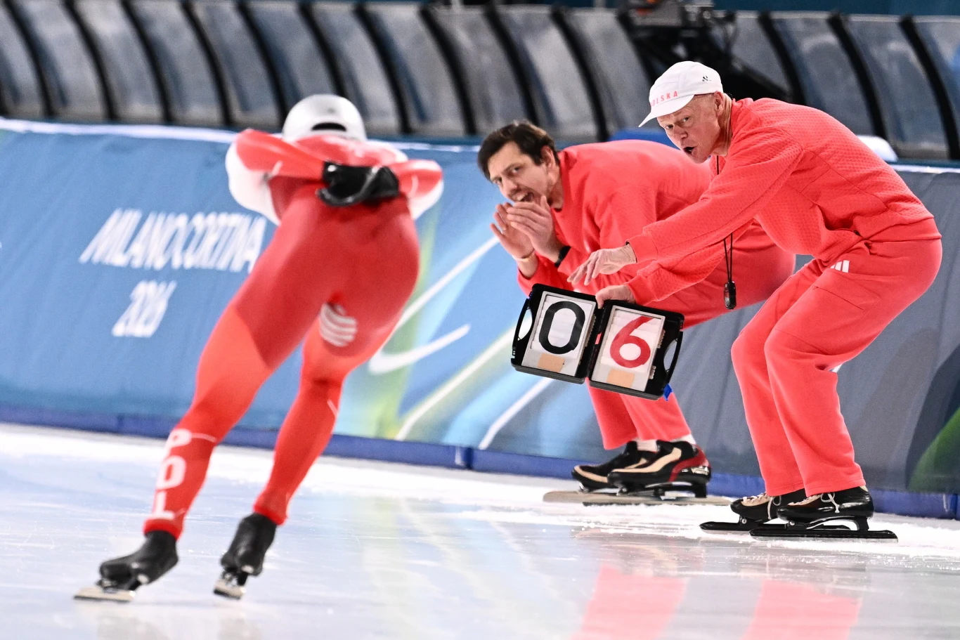 Trener Roland Cieślak dopinguje Władimira Semirunnija Zawodnik w czerwonym stroju łyżwiarskim ściga się na torze lodowym, a dwóch trenerów w identycznych uniformach przekazuje mu informacje liczbowe podczas rywalizacji.