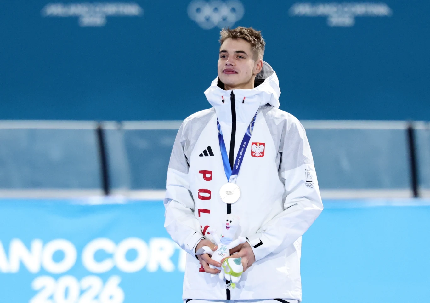 Młody sportowiec ubrany w biały strój z napisem Polska i godłem narodowym stoi na podium z zawieszonym na szyi srebrnym medalem, trzymając maskotkę imprezy. W tle widoczne są elementy dekoracji olimpijskiej oraz logo igrzysk zimowych Mediolan-Cortina 2...