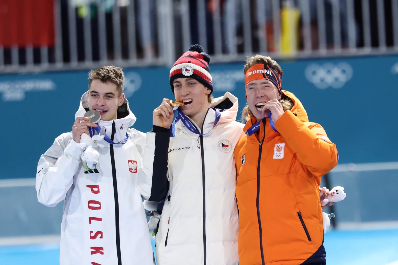 Trzech sportowców ubranych w stroje reprezentacyjne, każdy z medalem olimpijskim, gryzących swoje medale podczas dekoracji. W tle widoczne logo igrzysk olimpijskich oraz barierki oddzielające strefę dla zawodników.