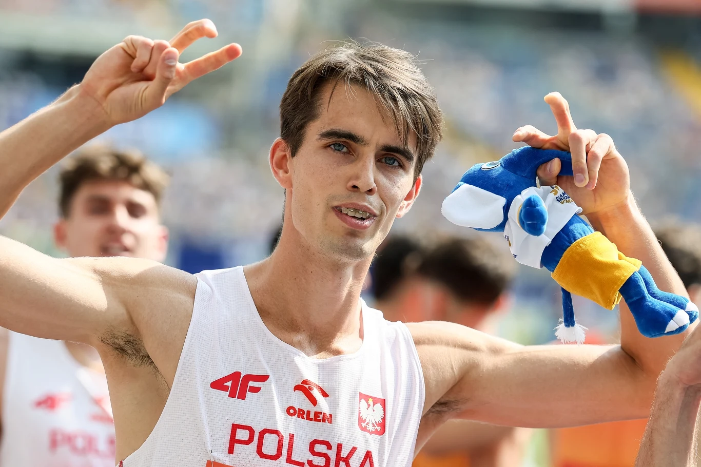 Kajetan Duszyński Sportowiec w białej koszulce z napisem 'Polska' wykonuje gesty zwycięstwa, trzymając pluszową maskotkę. W tle inne osoby, rozmyte oraz stadionowe trybuny.