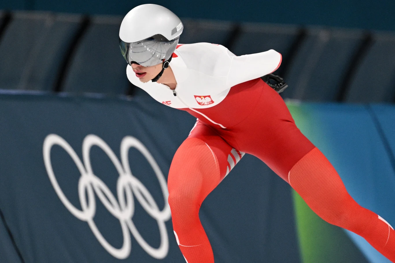 Władimir Semirunnij na igrzyskach olimpijskich w Mediolanie 2026 Łyżwiarz szybki w biało-czerwonym stroju z polską flagą ściga się na lodowisku olimpijskim, w tle znajdują się pierścienie olimpijskie.