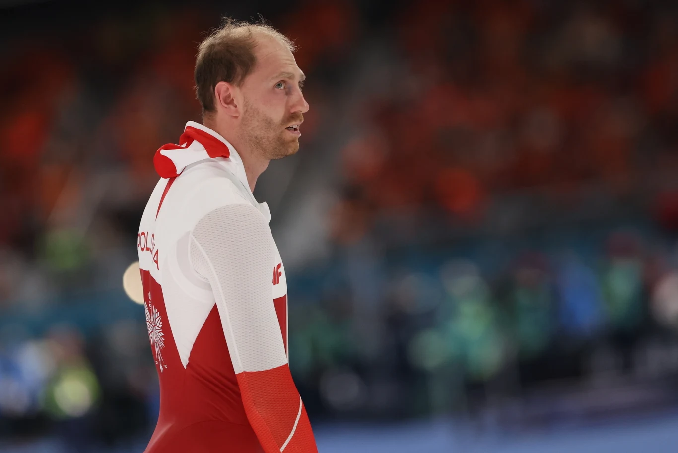 Sportowiec ubrany w biało-czerwony strój z napisem 'Polska' na plecach stoi na lodowisku, w tle rozmyci kibice i zawodnicy