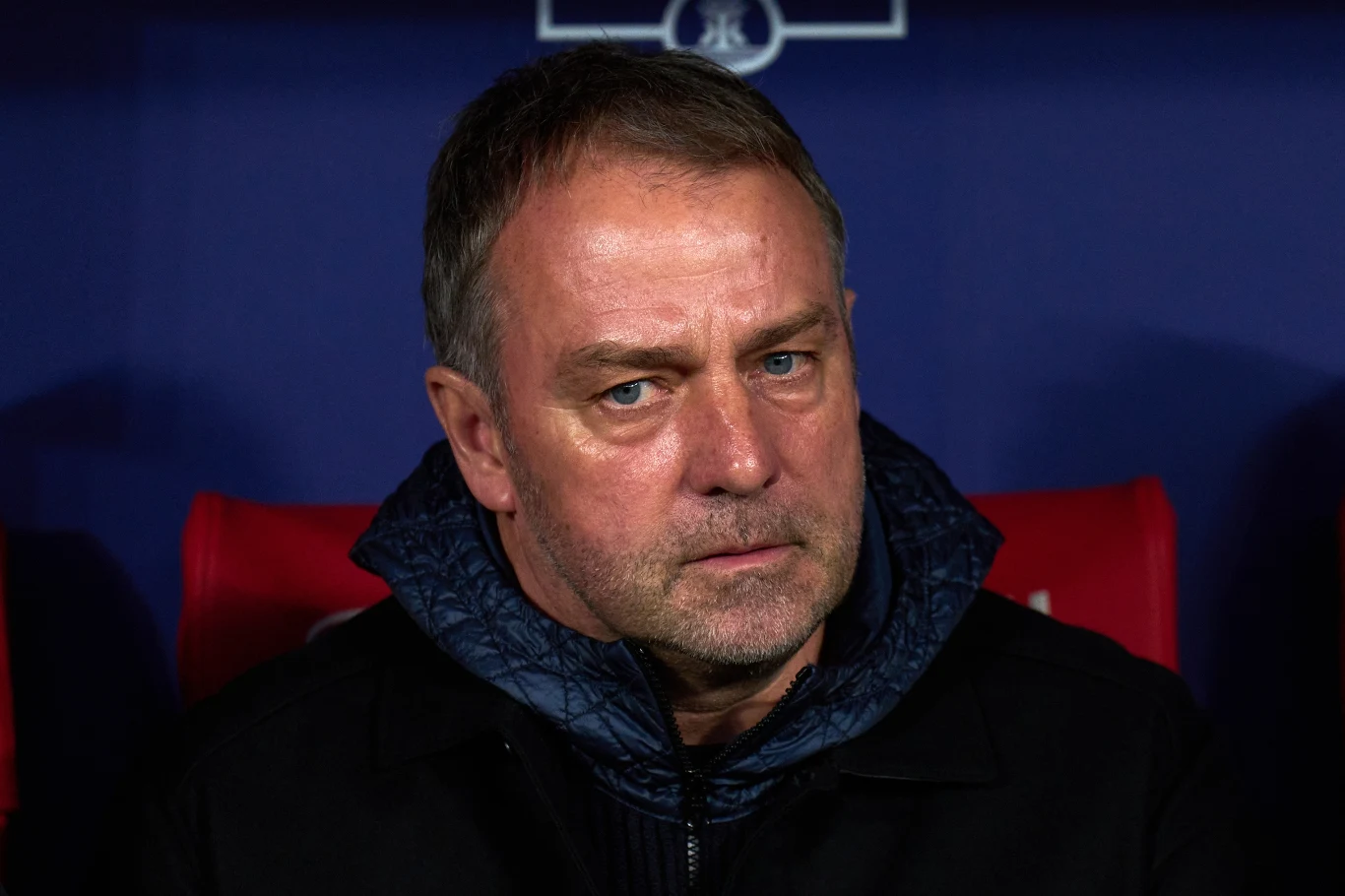 Hansi Flick Mężczyzna w średnim wieku w ciemnej kurtce z kapturem, siedzący na tle granatowej ściany z elementami czerwieni, patrzący wprost w obiektyw z poważnym wyrazem twarzy