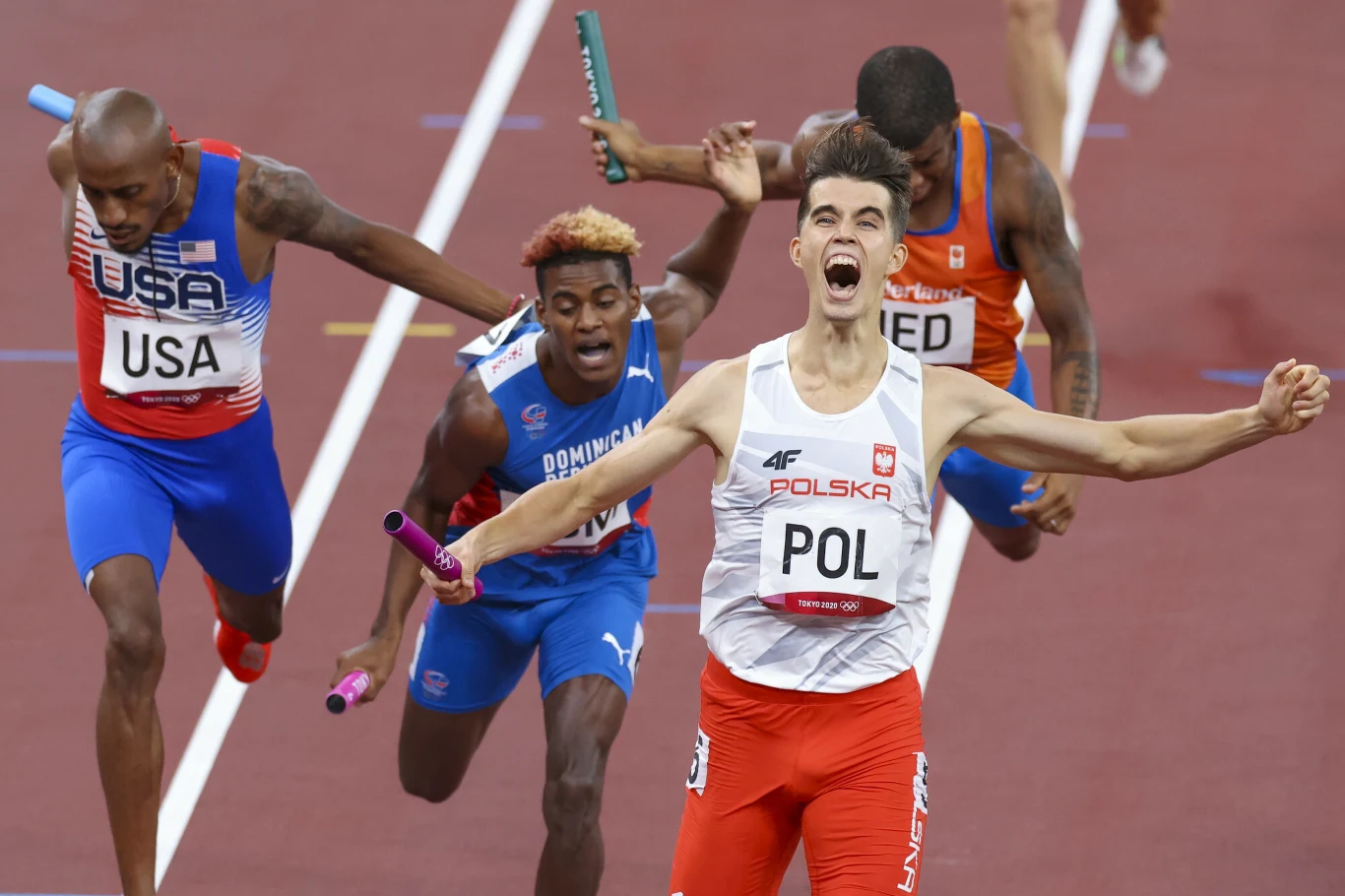 Kajetan Duszyński, Tokio 2021 Finisz sztafety lekkoatletycznej, sportowcy różnych narodowości walczą o zwycięstwo, polski zawodnik z wyciągniętymi ramionami świętuje triumf na mecie, widoczne emocje i determinacja uczestników.