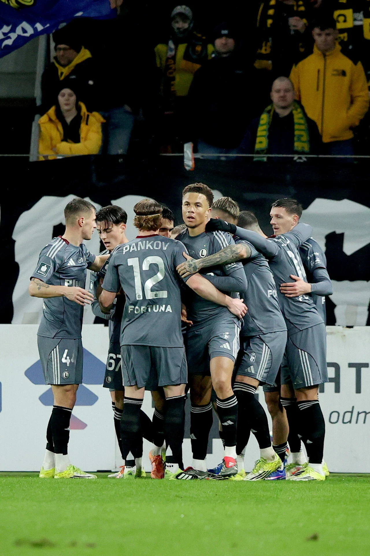 Po raz pierwszy w tym roku Legia wyszła na prowadzenie, ale pełnej puli z Katowic nie wywiozła Piłkarze w szarych strojach zgromadzeni w grupie świętują zdobycie gola, w tle kibice na trybunach ubrani na żółto-czarno.