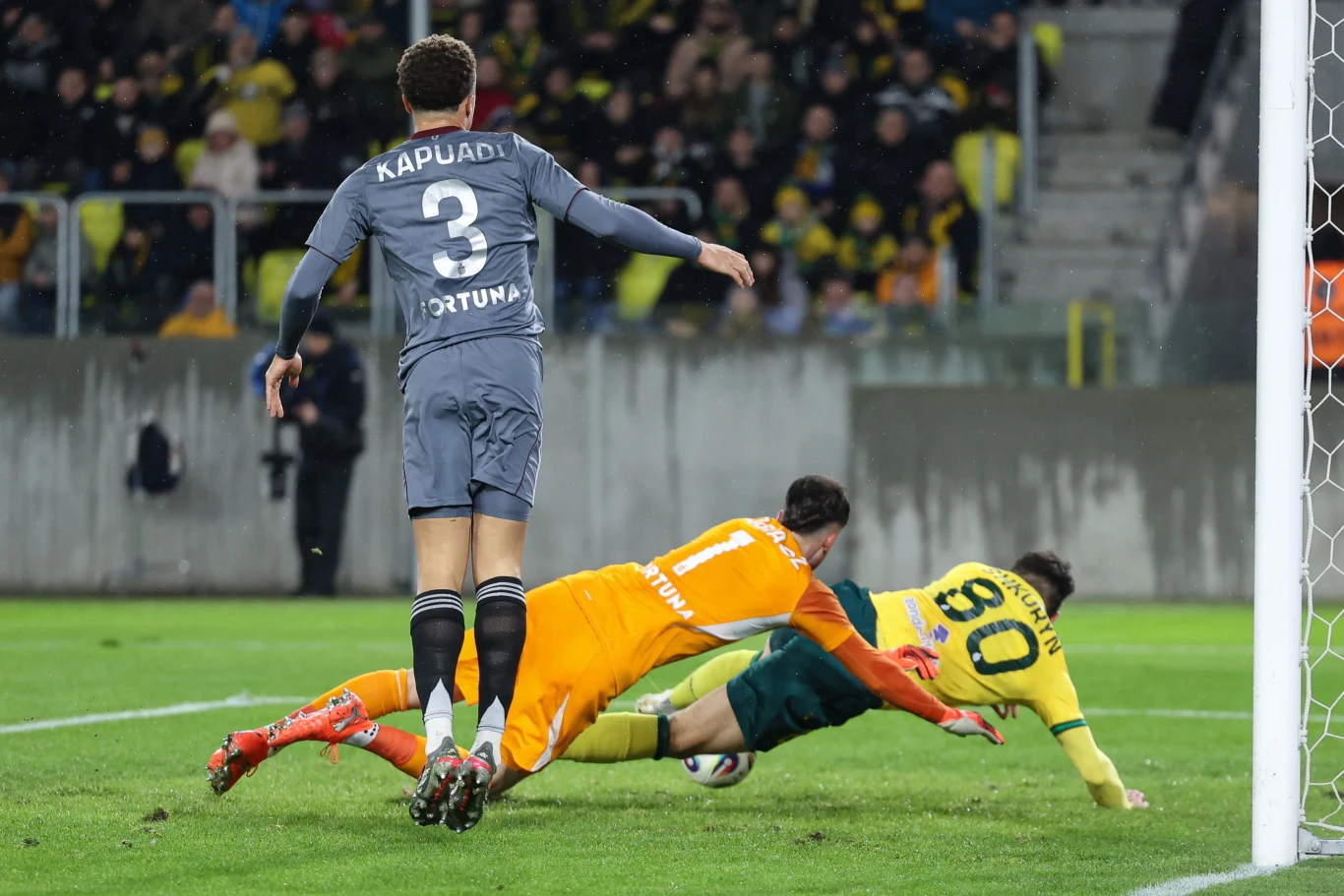 Po tej interwencji Kacpra Tobiasza potrzebna była analiza VAR. Wbrew pozorom Ilja Szkurin nie był faulowany Bramkarz i obrońca w pomarańczowych i szarych strojach próbują powstrzymać zawodnika w żółtej koszulce tuż przy bramce, dynamiczna akcja na tle trybun z kibicami.