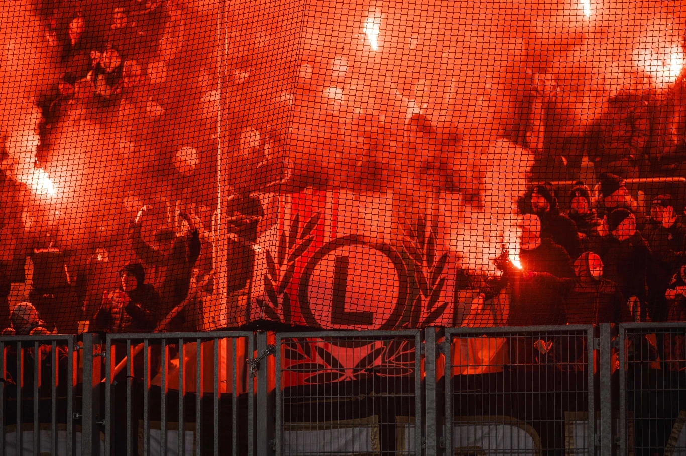 Kibice Legii prezentują się w tym sezonie zdecydowanie lepiej niż piłkarze Tłum kibiców z zapalonymi racami tworzy intensywną czerwoną poświatę, w centrum duża flaga z literą L na tle liści laurowych, przez siatkę stadionową widać uniesione ręce i płomienie.