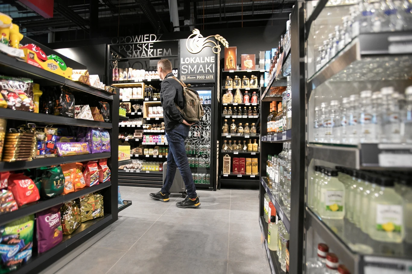 Mężczyzna z plecakiem przechodzi alejką sklepową pomiędzy regałami z przekąskami, napojami i alkoholem, otoczony produktami spożywczymi oraz lokalnymi wyrobami.