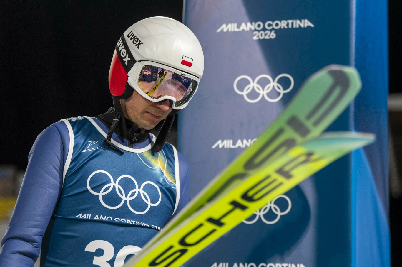 Kamil Stoch Skoczek narciarski w kasku i goglach z widocznym logo Olimpiady Milano Cortina 2026, skupiony przed oddaniem skoku, stojący przy nartach oraz oficjalnym banerze olimpijskim.