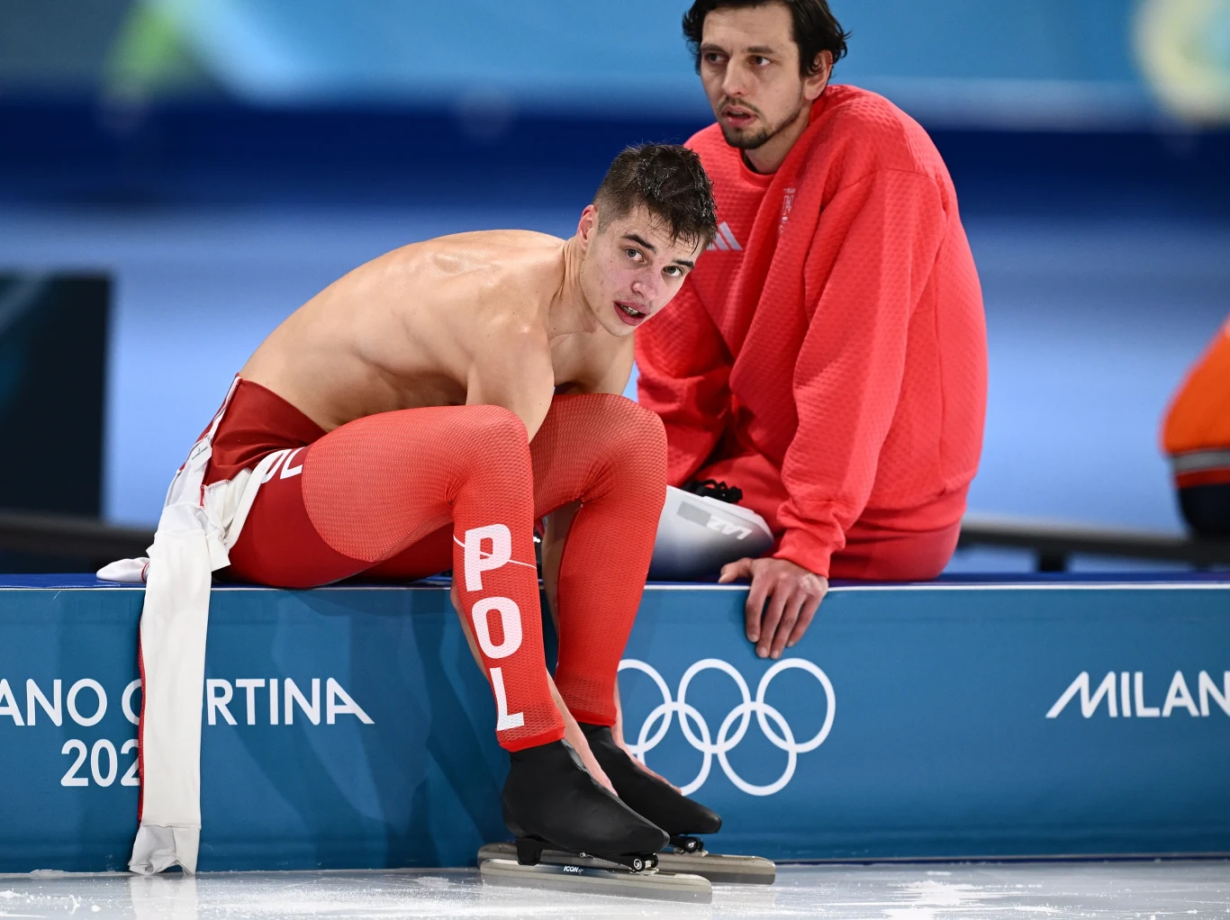 Władimir Semirunnij i trener Roland Cieślak Młody łyżwiarz szybki w czerwonym stroju z napisem POL siedzi na ławce przy lodowisku po zakończonym wyścigu. Obok stoi trener lub członek ekipy również w czerwonym ubraniu. Na niebieskiej barierce widnieje logo igrzysk olimpijskich Milano Cortina 2026.