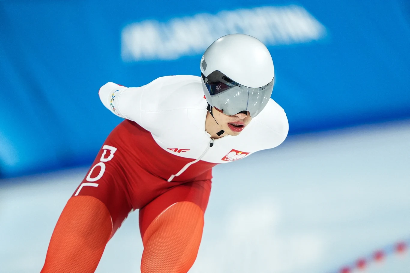 Łyżwiarz szybki w czerwono-białym stroju startowym z napisem POL oraz godłem Polski wykonuje dynamiczny ślizg na lodowisku, w tle rozmazane elementy niebieskiego toru.