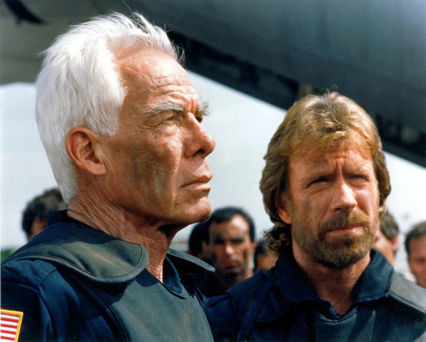 Lee Marvin i Chuck Norris w filmie "Oddział Delta" Dwóch mężczyzn w mundurach z lekko zabrudzonymi twarzami stoi na tle grupy osób, w tle elementy wojskowego sprzętu. Jeden z nich ma siwe włosy, drugi jasnobrązowe, obaj skoncentrowani i poważni.