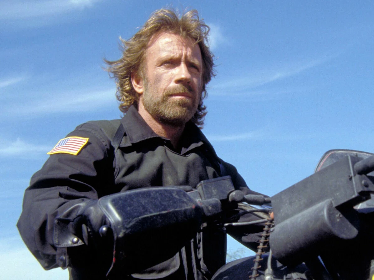 Chuck Norris w filmie "Oddział Delta" Mężczyzna z brodą i dłuższymi włosami ubrany w czarną kurtkę z naszywką flagi Stanów Zjednoczonych kieruje motocyklem na tle błękitnego nieba.