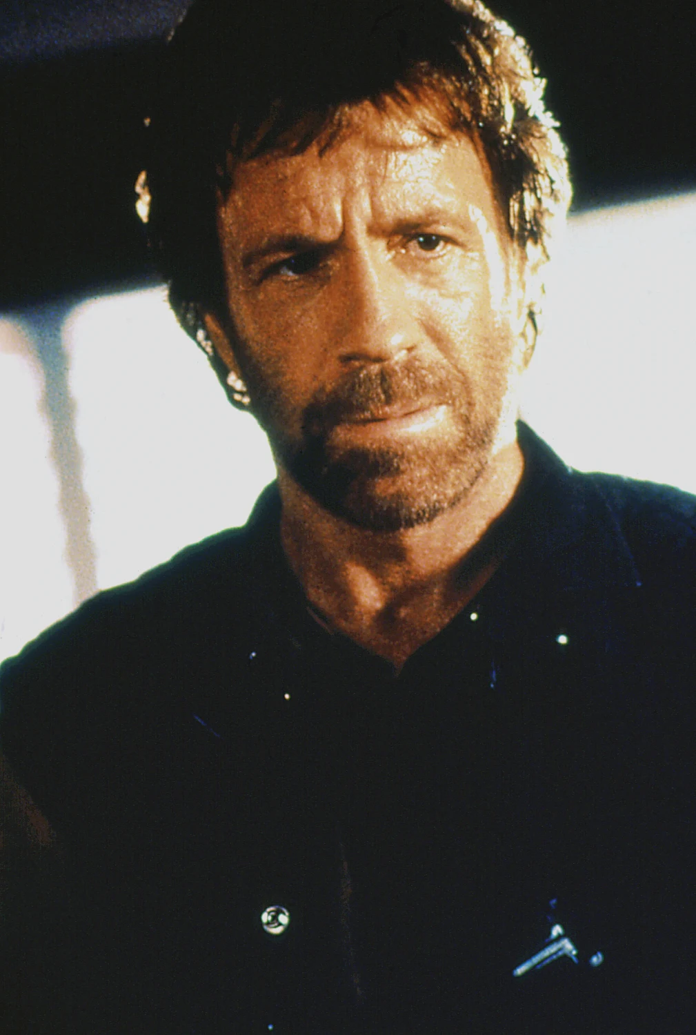 Chuck Norris w filmie "Oddział Delta" Mężczyzna o poważnym wyrazie twarzy z krótką brodą i burzą włosów, ubrany w ciemną koszulę, znajduje się na pierwszym planie na tle rozmytego, jasnego tła.