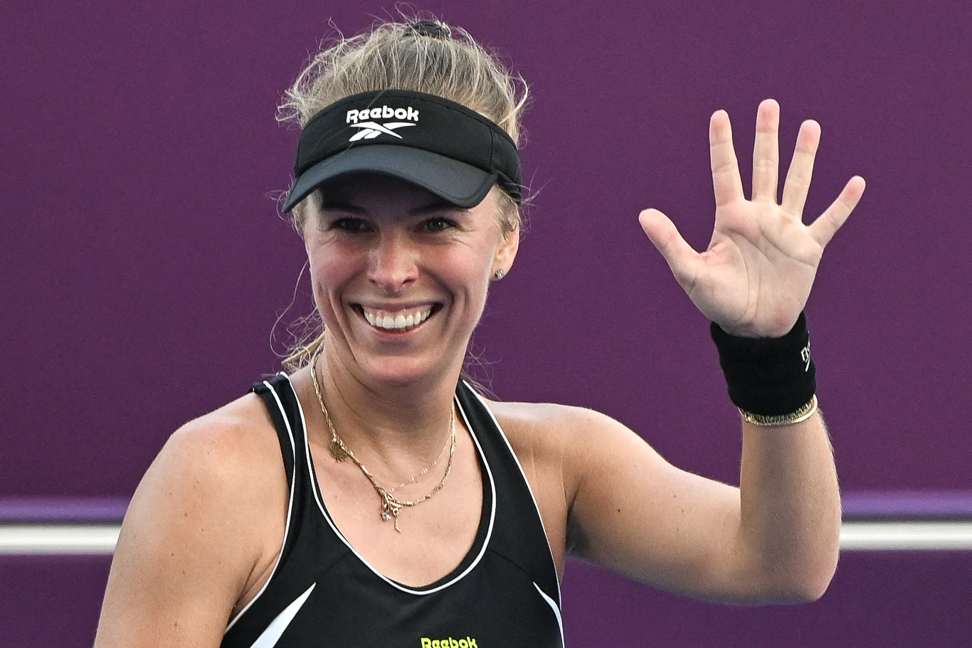 Uśmiechnięta kobieta w sportowej czarnej odzieży i czapce z daszkiem unosi dłoń w geście powitania na tle fioletowej ściany.