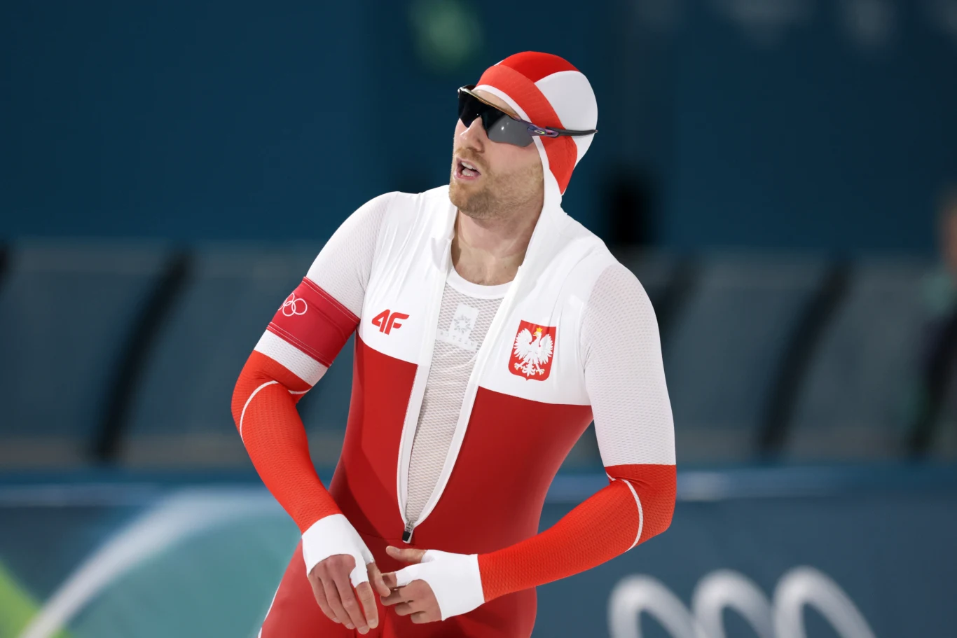 Sportowiec ubrany w biało-czerwony strój startowy z godłem Polski, noszący czapkę w tych samych barwach oraz okulary sportowe, stoi na lodowisku podczas zawodów.