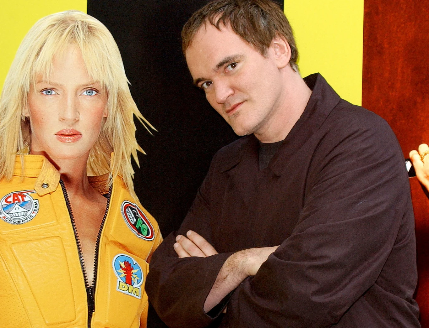 Quentin Tarantino na tle plakatu z Umą Thurman z filmu "Kill Bill : Volume 2" Mężczyzna ubrany w ciemną bluzę stoi z założonymi rękami obok dużego kartonowego wycięcia postaci kobiety w żółtym kombinezonie z naszywkami, na kolorowym tle.