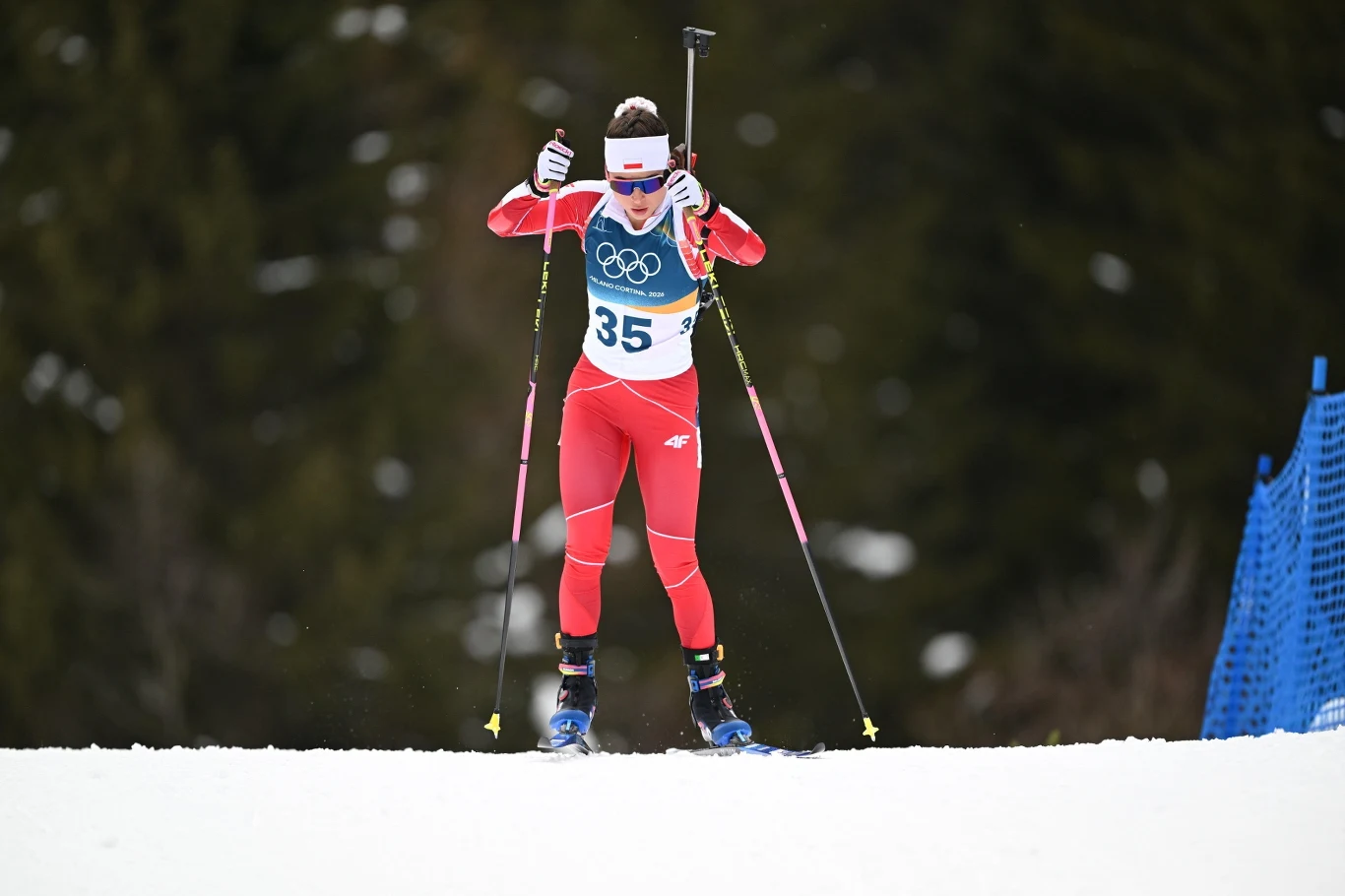 Kamila Żuk Zawodniczka w czerwonym stroju sportowym jedzie na nartach biegowych, trzymając kijki i karabin przewieszony przez plecy, na tle zaśnieżonego terenu i ciemnych drzew, bierze udział w zawodach olimpijskich.