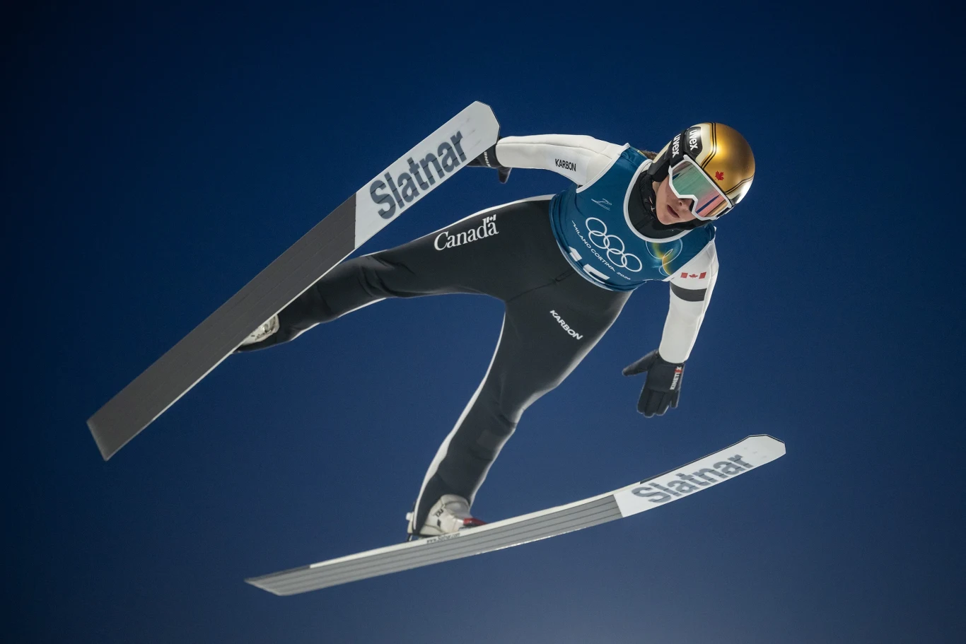 Abigail Strate Skoczek narciarski w locie podczas zawodów, ujęty na tle bezchmurnego nieba, w stroju sportowym z charakterystycznym kaskiem i goglami, numerem startowym oraz widocznymi nartami z nazwą sponsora.