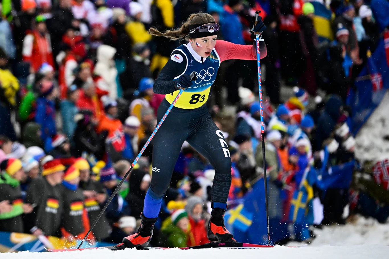 Zawodniczka w stroju narciarskim bierze udział w wyścigu biegów narciarskich na tle kibicującego tłumu, wśród którego widoczne są liczne flagi narodowe, w tym szwedzkie.