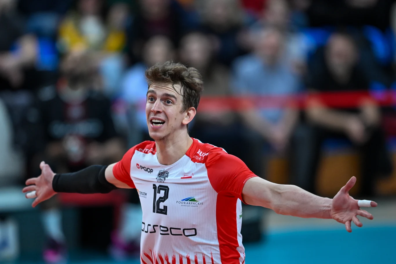 Siatkarz w biało-czerwonym stroju z rozłożonymi rękami, stojący na hali sportowej z rozmytym tłem pełnym widzów.