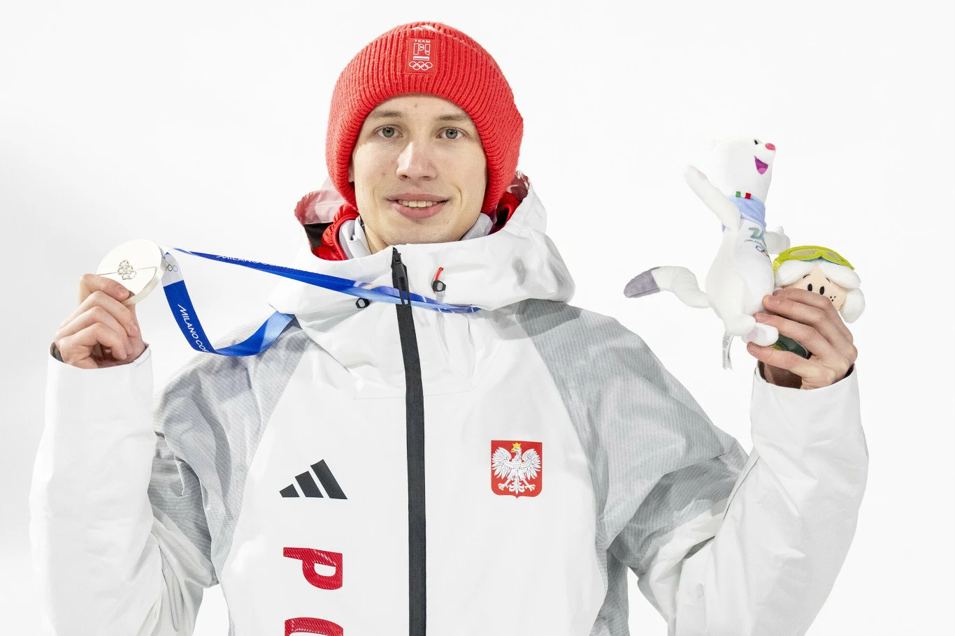 Kacper Tomasiak ze srebrnym medalem Sportowiec w biało-czerwonym stroju z godłem Polski na piersi, trzyma w jednej ręce medal na niebieskiej wstążce, a w drugiej maskotkę, ubrany w czerwony zimowy czepek, na białym tle.