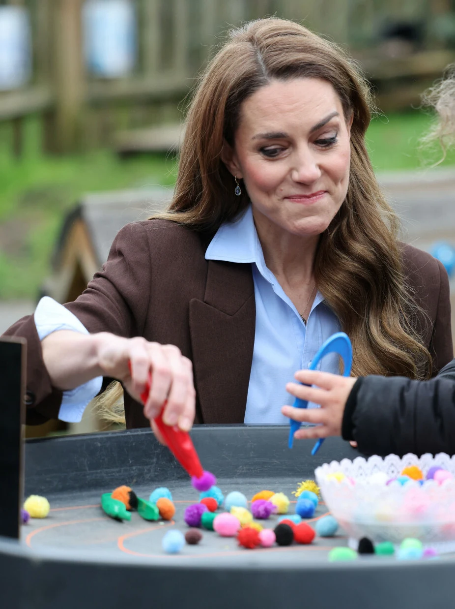 Kobieta w eleganckiej marynarce o długich, brązowych włosach siedzi przy okrągłym stole z różnokolorowymi pomponami i narzędziami do zabawy, obok niej znajduje się dziecko uczestniczące w zabawie rękami.