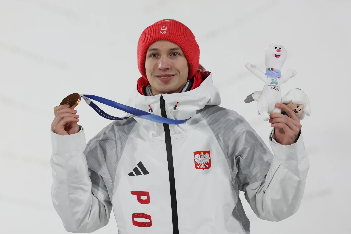 Sportowiec ubrany w biały strój z polskim godłem na piersi trzyma brązowy medal olimpijski i maskotkę igrzysk zimowych. Na głowie czerwona czapka, w tle neutralne, białe otoczenie.