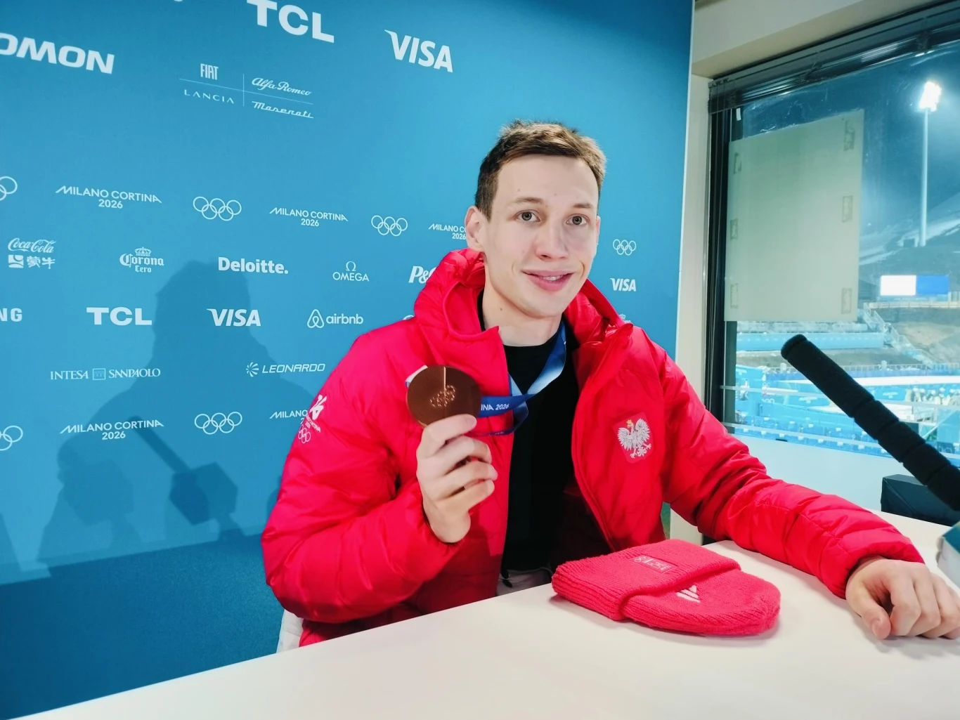 Sportowiec w czerwonej kurtce z emblematem Polski siedzi przy stoliku, trzymając w ręku brązowy medal na tle ściany z logo sponsorów i zimowego obiektu sportowego. Na stole leżą czerwone rękawiczki.