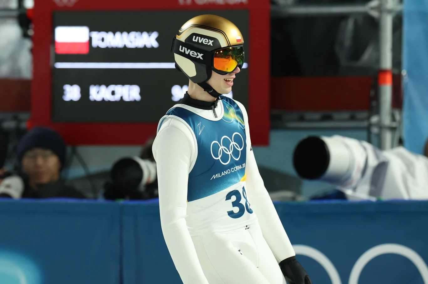 Zawodnik w kombinezonie narciarskim i złotym kasku z goglami na tle tablicy wyników podczas zimowej olimpiady.